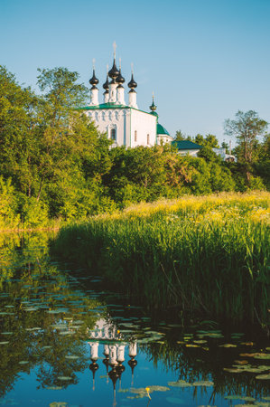 Orthodox church with black houses reflects in a calm river surrounded by summer greeneryの写真素材