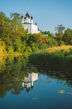 Orthodox church with black houses reflects in a calm river surrounded by summer greeneryの写真素材