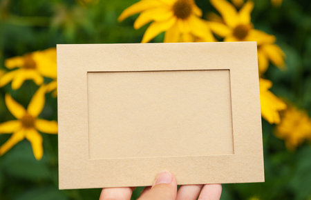 Hand of woman holds the empty beige paper against green leaves and flowers natural backgroundの写真素材