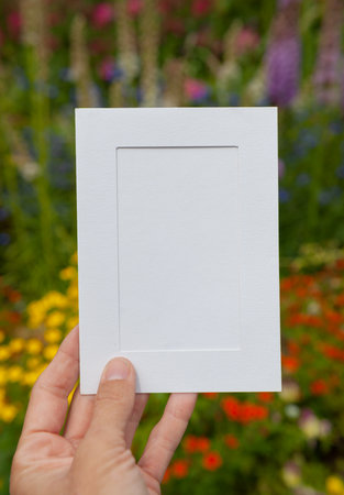 Hand of woman holds the empty white paper against green leaves and flowers natural background