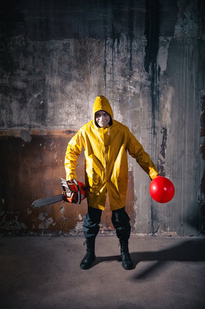 Sinister man in yellow raincoat holds red balloon and chainsaw in grungy basement evokes Halloween horror vibeの写真素材