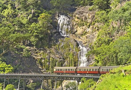 Kuranda scenic railway winling up the tracks from Freshwater Station to Kuranda. The train passes by a long bridge and a nice waterfal behindの写真素材