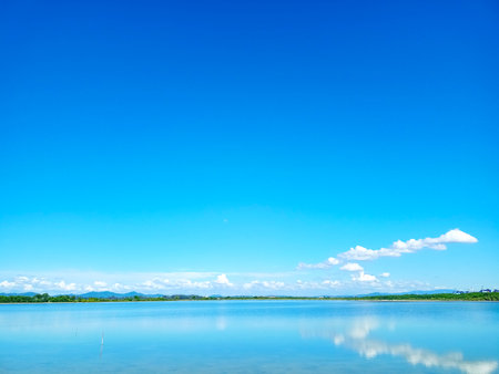 reflection of blue sky and white cloud on lake surface with line of tree and tiny hillの写真素材