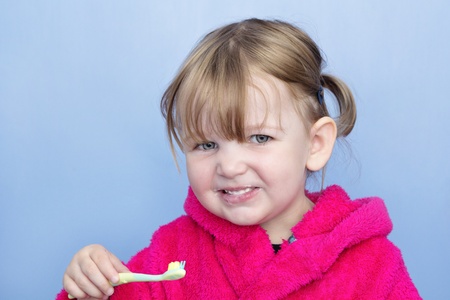 A young girl in a pink dressing gown against a light blue background. She's cleaning her teeth and smiling at the camera.の写真素材
