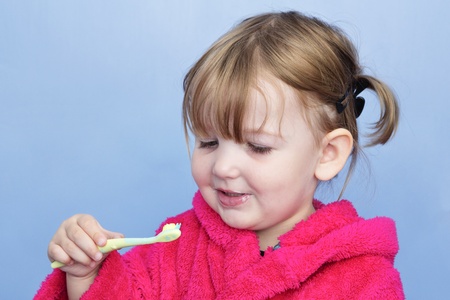 A young girl in a pink dressing gown against a light blue background. She's cleaning her teeth and has stopped to smile at the brush.の写真素材