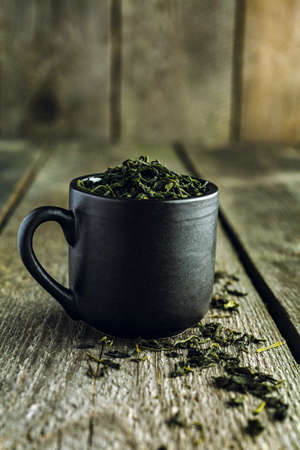 Dry organic green tea leafs in a black cup on a wooden backgroundの写真素材