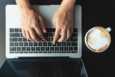 Hands typing a button on the laptop on a black wooden table in the cafe in the morning. And latte coffee in a white coffee mug beside, top viewの写真素材