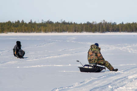 Winter fishing. Ice fisherman fishing in the winter on the river. winter fisherman sits in camouflage clothing near a hole with an ice drill on a frozen snow covered river on a winter day.の写真素材
