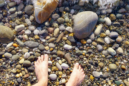 Close up view of a man feet into the pebbles on the beach with sea water flowingの写真素材