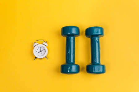 Dumbbell, alarm clock on yellow background. Minimalistic sport concept. Top view.