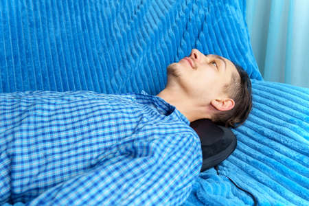 Man lies on the couch and massage his neck with a electric massage pillow.