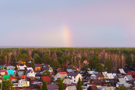 Colorful rainbow arcs over a tranquil village surrounded by lush green trees after a gentle rain in early springの写真素材