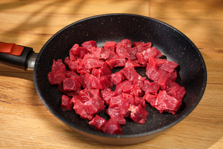 Chunks of raw beef sit in a black skillet on a wooden countertop. Bright light casts soft shadowsの写真素材