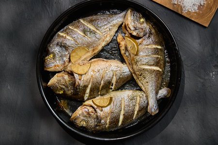 Grilled dorado with lemon slices arranged on a black serving dish garnished with herbs