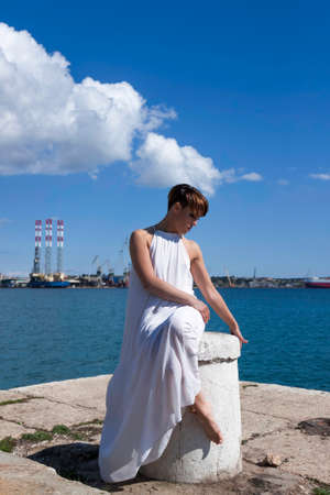 Female sitting on pier in white dress with blue skies and sea in background.の写真素材