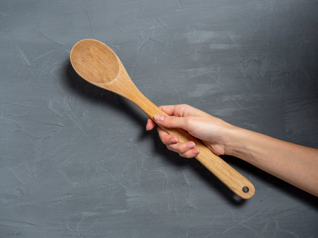 A woman's hand holds a large wooden spoon on a gray textured background. Tools for cooking food.の写真素材