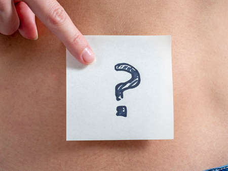 A sticker with a question mark is pasted on the bare female stomach. She points to the sticker with her finger. The concept of pregnancy and motherhood.の写真素材