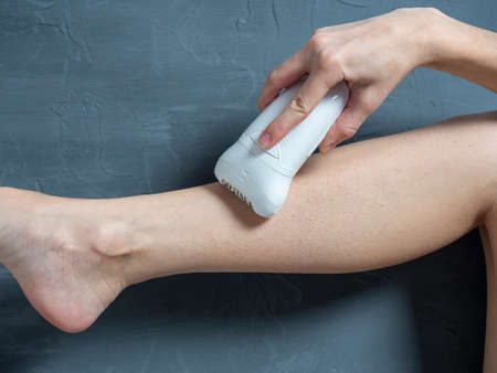 a woman shaves her leg with an electric epilator on a gray textured background. The concept of...