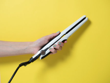 White curling iron in a woman's hand on a yellow background. An accessory for creating hairstyles. Beauty and fashion. Hair care.の写真素材