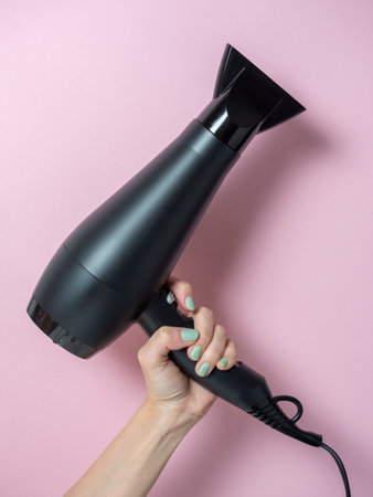 black hair dryer in a woman's hand on a pink background. Hair Care Product. Vertical photoの写真素材