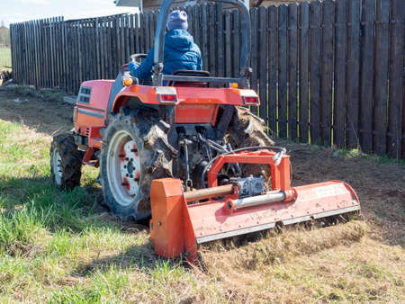 The man sits in a small tractor and drives through the field mulching grass. Land cultivation, agrotematicsの写真素材