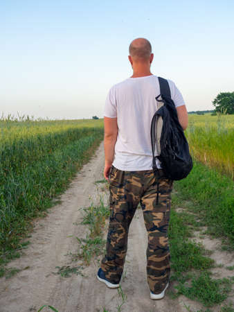 a man is standing on a path in a field with a black backpack with his back to the camera. Evening landscape, travel conceptの写真素材