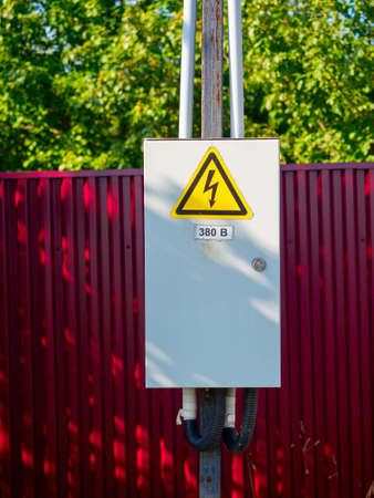 Close-up of a gray metal electric shield hanging on a pole. A warning sign. Red fence in the backgroundの写真素材
