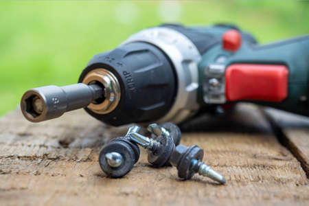 A close-up of an electric screwdriver with a nozzle on lies on a wooden background. Next to it, roofing screws on metal are laid out in a row. The concept of repair and constructionの写真素材