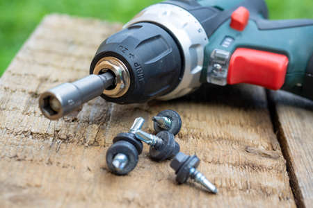 A close-up of an electric screwdriver with a nozzle on lies on a wooden background. Next to it, roofing screws on metal are laid out in a row. The concept of repair and constructionの写真素材
