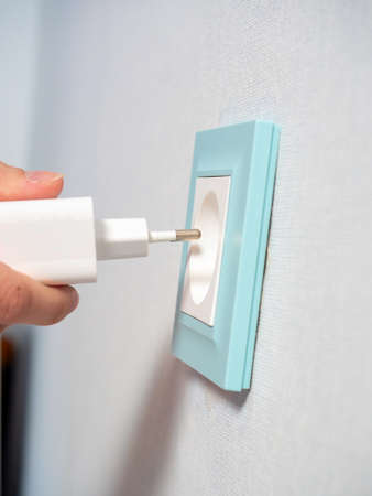 Close-up of a woman's hand holding a white plug and about to plug it into a blue outlet on the wall in the house. The concept of the need for electricity in the house. Mobile devices, Electrificationの写真素材