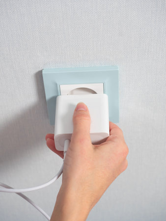 Close-up of a woman's hand holding a white plug and about to plug it into a blue outlet on the wall...