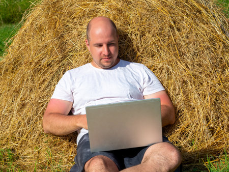 a middle-aged man, dressed in a T-shirt and shorts, is lying on a yellow haystack with a gray laptop in his hands. Remote work, training. Rural area.の写真素材