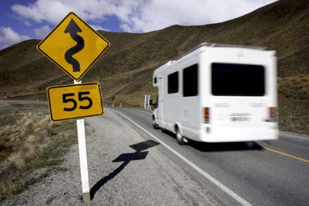 White campervan/RV heading downhill at speed in New Zealand with a winding road warning sign. の写真素材