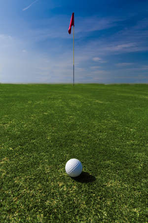 Close up of a ball on a green with a flag in the distance against a blue skyの写真素材