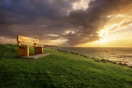 A bench on a seaside cliff with a dramatic sunrise loooking out over the ocean as the storm clouds...
