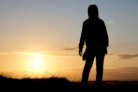 A low-angle view of a silhouetted woman stood on a ridge in the countryside as the bright summer sun sets to the sideの写真素材