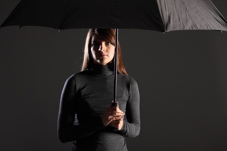 Beautiful young woman standing under the protection and cover of an umbrella. Model wearing dark clothing, looking straight into camera. Dramatic lighting to emphasise danger and need for shelter.の写真素材
