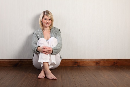 Beautiful young caucasian woman with blonde hair and happy smile sitting on laminate floor at home, relaxing bare foot. She is wearing white linen pants and a grey heavy knit cardigan.の写真素材