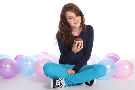 Pretty teenager girl celebrates happy birthday with a chocolate cup cake and single pink candle, sitting among party balloons with a happy smile.の写真素材
