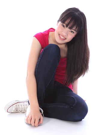 Sitting on the floor a beautiful young Japanese teenager high school student girl, with long black hair wearing blue denim jeans and a pink t-shirt with big happy smile.の写真素材