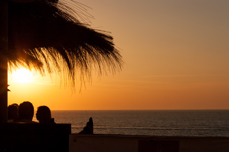 silhouette of people at sunset under a straw umbrellaの写真素材