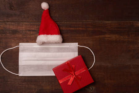 Protective mask and Santas cap with gift box, flat lay with space for text on brown wooden backgroundの写真素材