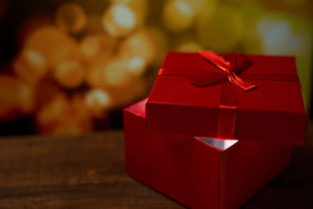 Red gift with a bow and with light inside on a brown wooden table on a gold bokeh background with a copy spaceの写真素材