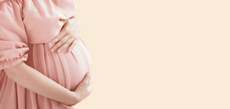 Pregnant woman in pink dress hugs her belly on beige background. High quality photo