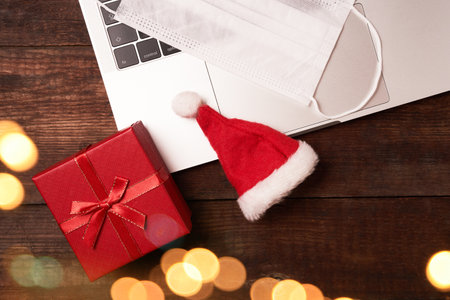 Online safe shopping for Christmas, a laptop stands on a wooden table next gift box, a Christmas cap and face maskの写真素材