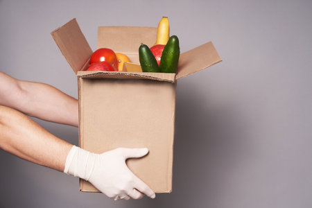Hands of white courier in rubber gloves hold a cardboard box of groceries, fast and safe food...