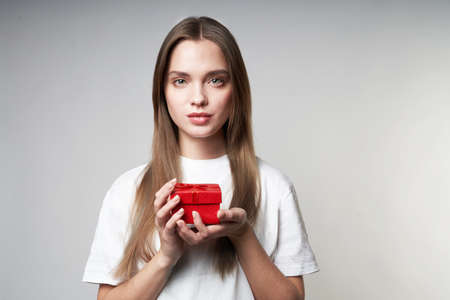 Beautiful young blonde woman on a gray background with a red small gift in hands. High quality photoの写真素材