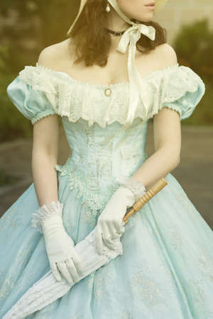 A beautiful young woman in a historical blue dress with an umbrella, gloves and a hat. Dress on a woman of the 19th century. Design for the book coverの写真素材