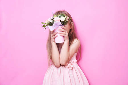 A little girl in pink dress with bouquet of flowers in a paper cup, hid behind the flowers. The girl is shy and sniffs the flowers on pink background.の写真素材