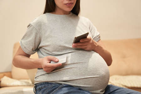 Pregnant woman at home with phone is sitting with credit cart, concept: online shopping. High quality photoの写真素材
